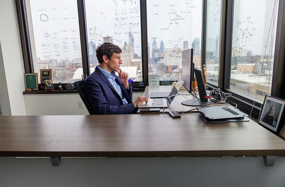 David Fajgenbaum sits at his desk and stares at a computer screen with the city skyline in the window behind him